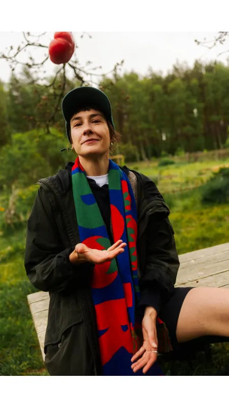 A person in a dark jacket, baseball cap, and colourful scarf sits outdoors on a wooden surface amid greenery, tossing an organic tomato—perhaps after exploring curated Tallinn shopping or visiting a local gift shop. Gate21.ee