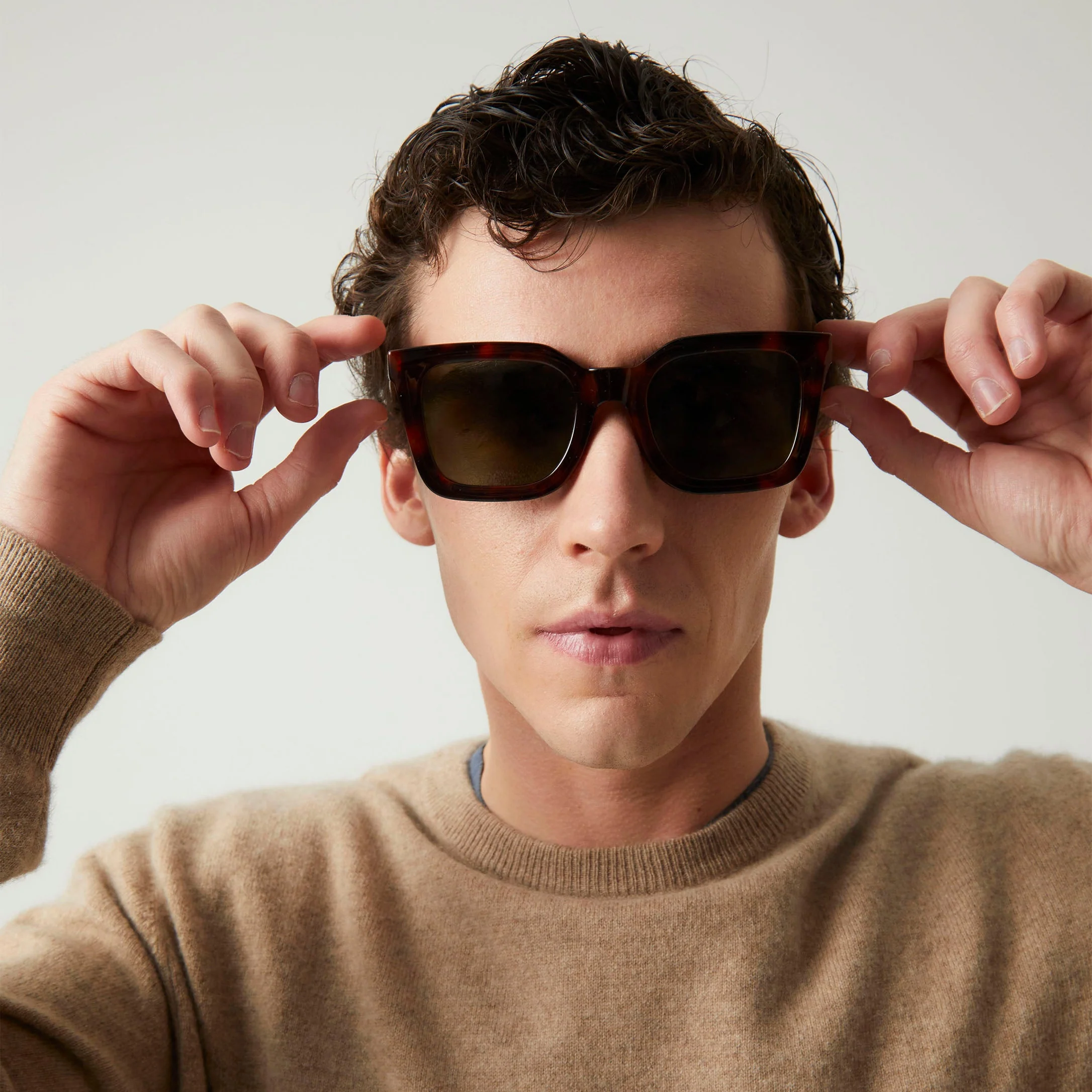 A person with short curly hair wears a tan jumper and adjusts the bold "Cometa" Turtle Sunglasses against a simple light background, showcasing the distinctive style found in concept shops that champion unique ideas. Gate21.ee