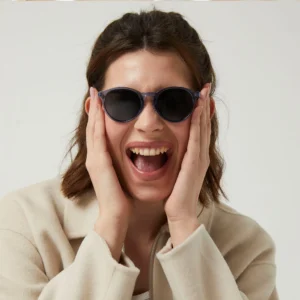 A person with shoulder-length brown hair wears "Isla" Blue Sunglasses and a beige jacket, holding their face in surprise at an Estonian design concept shop against a plain background. Gate21.ee