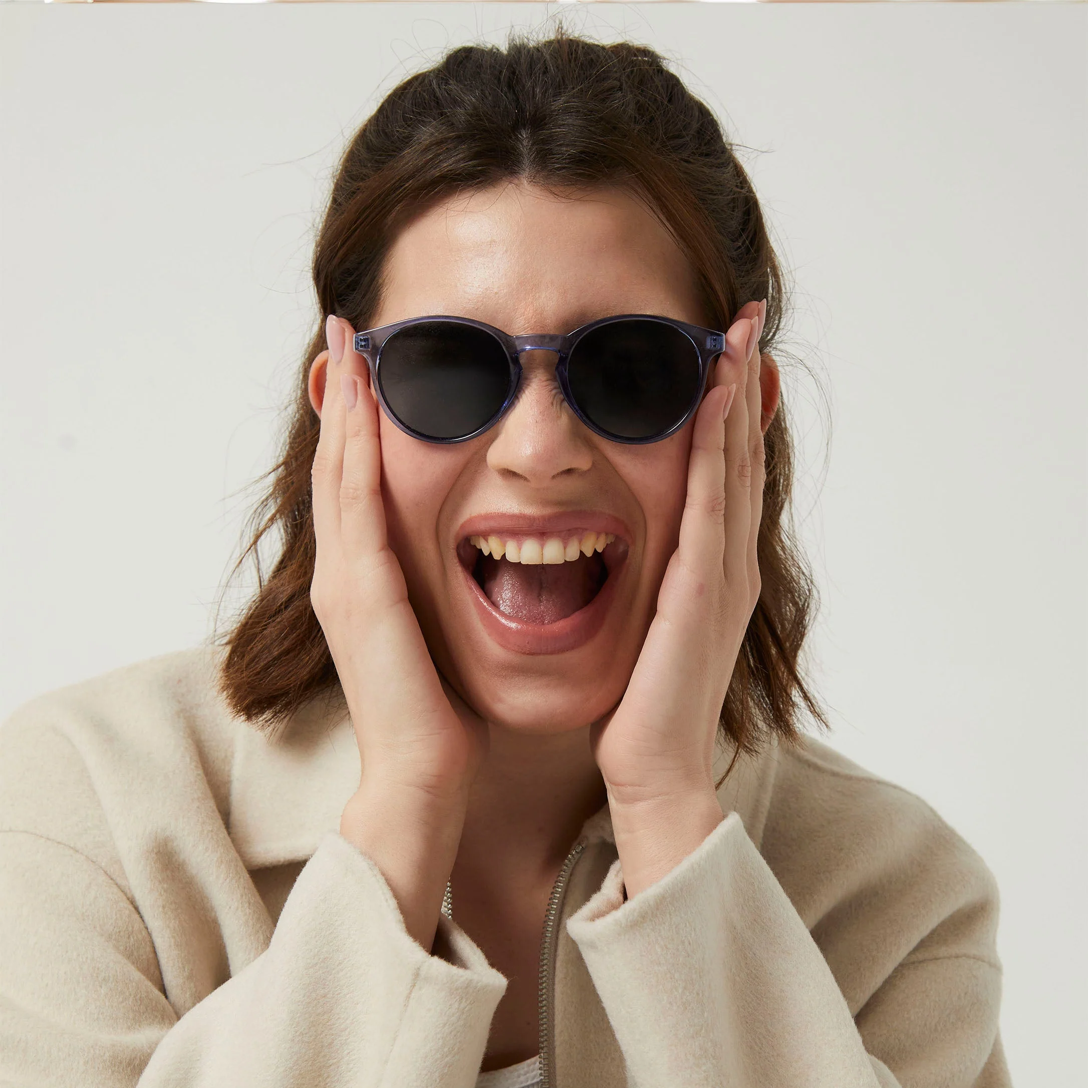 A person with shoulder-length brown hair wears "Isla" Blue Sunglasses and a beige jacket, holding their face in surprise at an Estonian design concept shop against a plain background. Gate21.ee