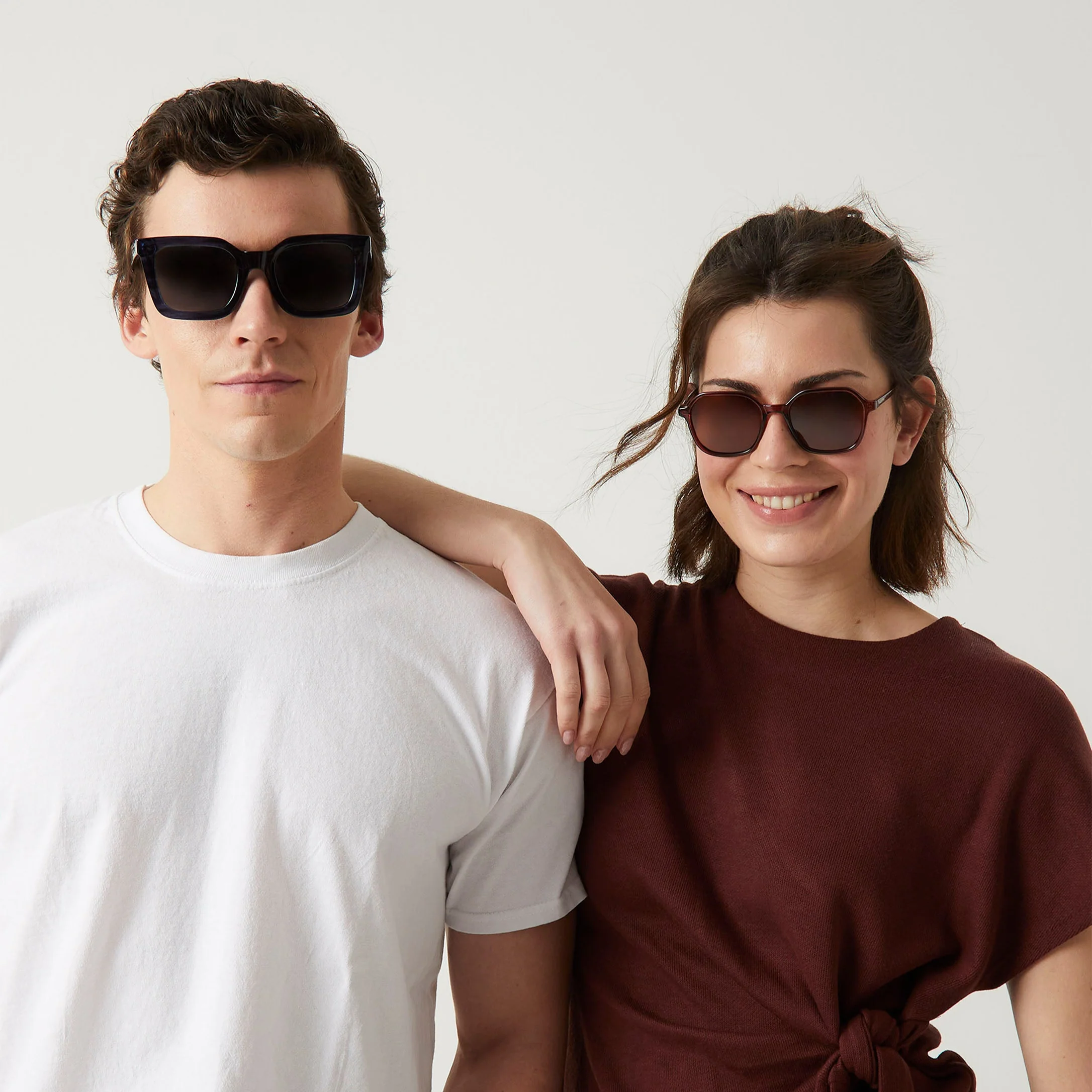Two people pose against a plain background as if for a gift shop editorial. One wears a white T-shirt with "Cometa" Deep Blue Sunglasses; the other, in a maroon top, rests their arm on their companion's shoulder. Gate21.ee