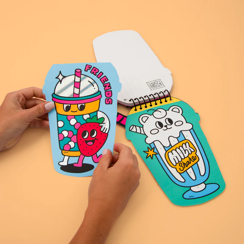 Two hands hold the "Bubble Tea - Sticker by Number & Postcards," a colourful, cup-shaped notepad with cartoon drink characters. Another shows a white bear and "Milk Shake," both featuring playful Estonian design on a light peach background. Gate21.ee