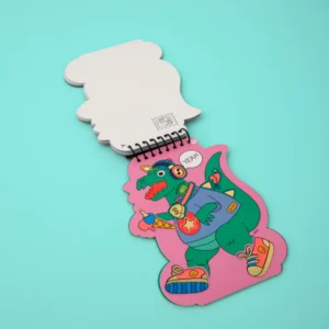 The Dino - Sticker Activity Book & Postcards features playful cartoon dinosaurs in fun outfits on a pink background, making it a vibrant and curated Estonian design perfect for adding colour to any gift shop display. Gate21.ee