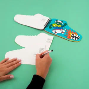 Someone fills in a Street - Sticker by Number & Postcards set shaped like trainers. Nearby are more trainer-shaped notecards and a notepad with a cartoon cat in sunglasses and &ldquo;COOL CAT&rdquo;&mdash;unique, concept shop-worthy gift items. Gate21.ee