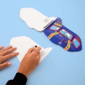 Someone is drawing on a page from Vroom – Sticker Activity Book & Postcards, a unique and colourful spiral-bound notepad, open on a light blue surface—perfect for concept store browsing or shopping in Tallinn. Gate21.ee
