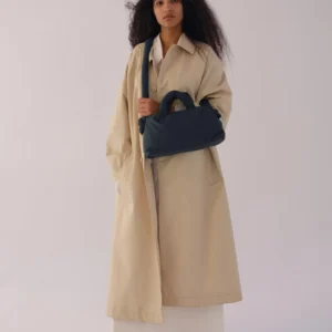 A person with long, curly hair stands barefoot on a white floor in a beige trench coat and white outfit, holding the Mini Ona Soft Bag - Coal—perfect for Tallinn shopping or discovering unique finds at a stylish gift shop. The background is plain and bright. Gate21.ee