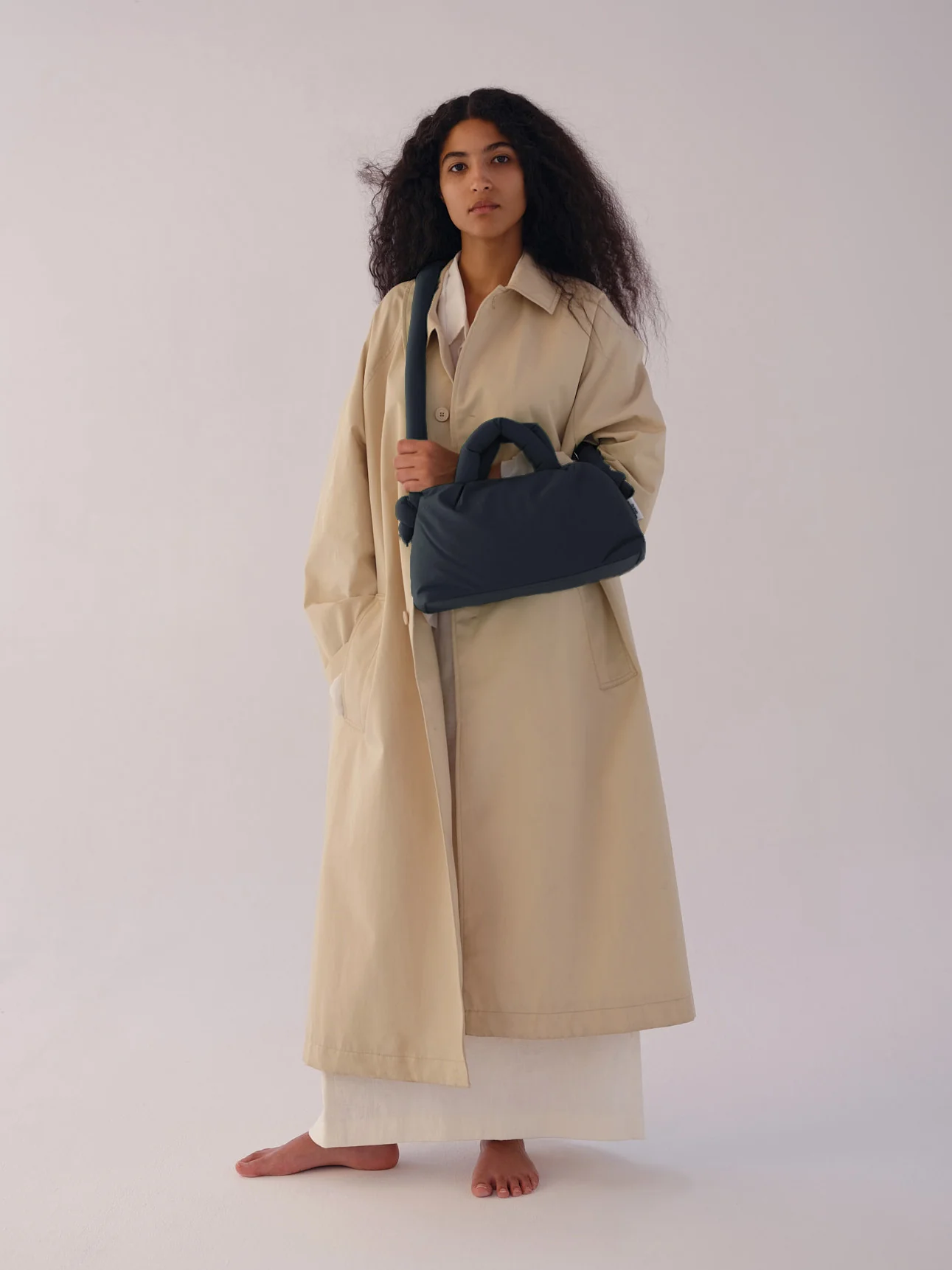 A person with long, curly hair stands barefoot on a white floor in a beige trench coat and white outfit, holding the Mini Ona Soft Bag - Coal—perfect for Tallinn shopping or discovering unique finds at a stylish gift shop. The background is plain and bright. Gate21.ee