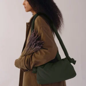 A person with long curly hair in a brown coat stands in profile against a plain backdrop, holding dried lavender—ideal for curated gift shops—and carrying the Mini Ona Soft Bag in Forest Green over their shoulder. Gate21.ee
