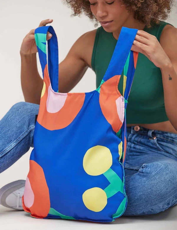 A person in a green sleeveless top and blue jeans holds the Blossom - Special Edition blue reusable bag with a colourful abstract pattern—ideal for shopping in Tallinn or discovering unique Estonian design at local concept shops. Gate21.ee
