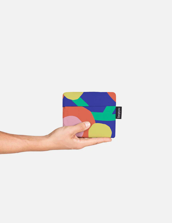A hand holds the Blossom – Special Edition pouch, a small square accessory with a vibrant abstract pattern—ideal for unique gift shops. It features a black tag with white text on the right, set against a plain white background. Gate21.ee