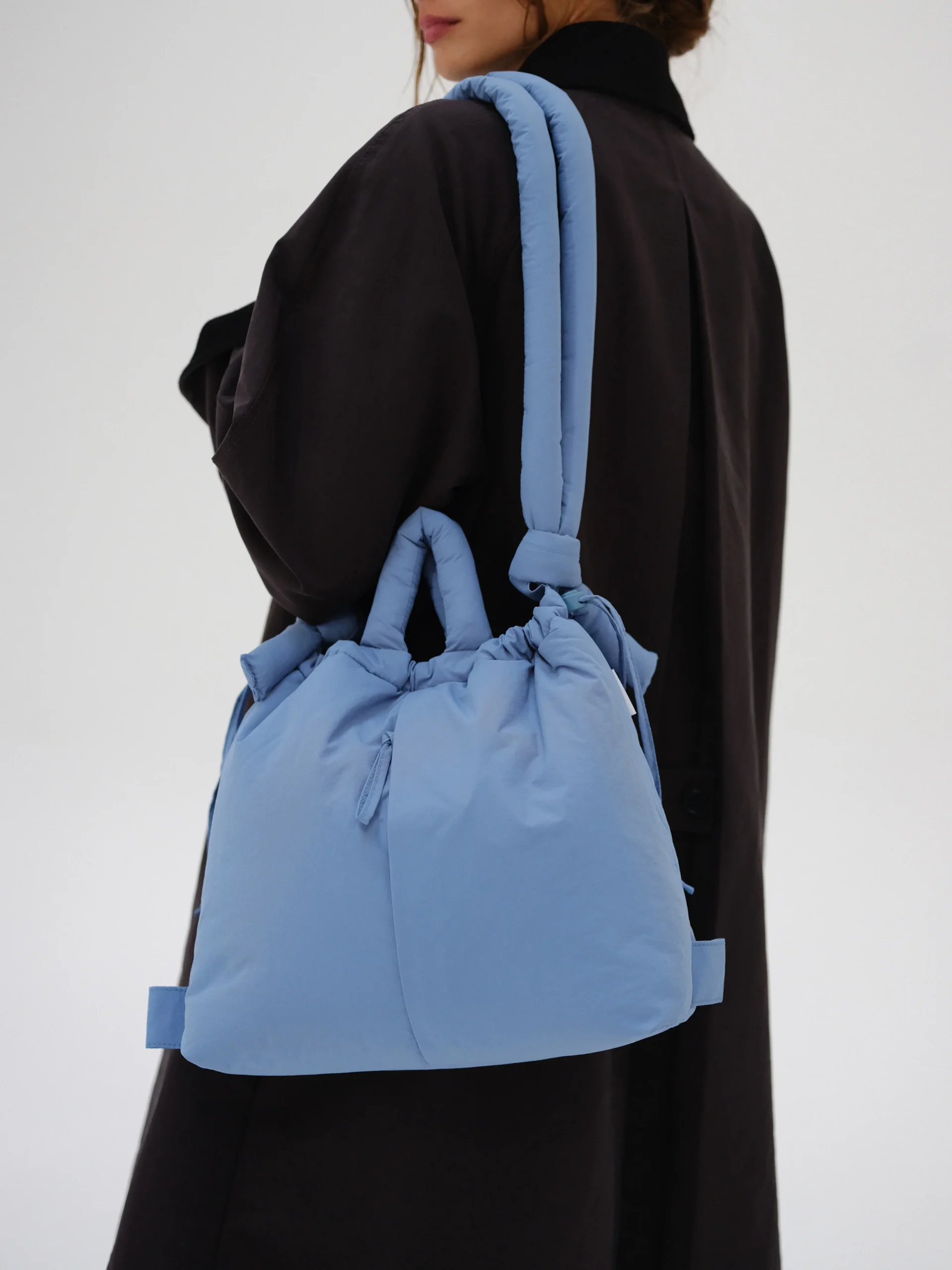 A person in a long, dark coat carries the Ona Soft Bag - Light Blue, a spacious padded bag with shoulder and hand straps—ideal for shopping in Tallinn or highlighting distinctive Estonian design—set against a plain, light background. Gate21.ee