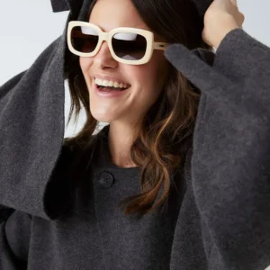A woman in a dark grey coat and "Duna" Creamy Sunglasses smiles with raised hands, radiating curated style—perfect inspiration for unique finds whilst shopping in Tallinn. Gate21.ee