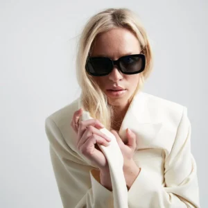 Wearing long blonde hair and a white blazer, a person in "Duna" Black Sunglasses stands against a plain light background, showcasing the modern style found in Tallinn's shopping concept stores. Gate21.ee
