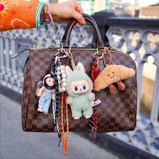 A brown chequered handbag with a dark handle is decorated with curated, colourful keyrings&mdash;a doll, plush bunny, and smiling croissant. A person wearing bracelets holds the bag outdoors near a bridge railing, perfect for Tallinn shopping. Gate21.ee