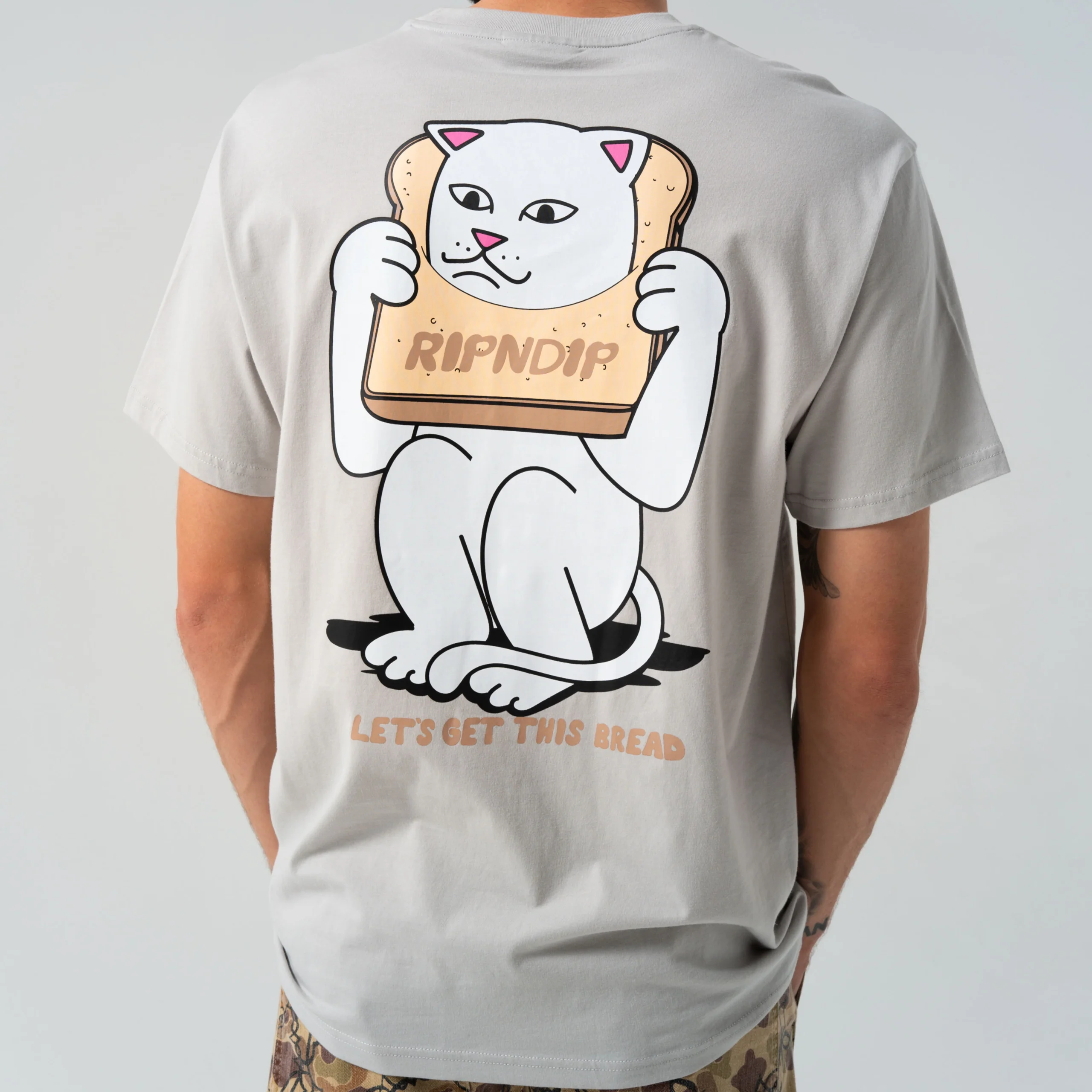A person in the Gluten Free Tee Grey—a light grey shirt with a white cat holding bread, "RIPNDIP" and "LET'S GET THIS BREAD" print—perfect for Tallinn shopping or anyone seeking unique, curated streetwear. Gate21.ee