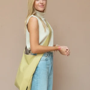 A person with long blonde hair stands against a beige wall in a sleeveless beige top and light blue jeans, carrying the Feel Good Bag - Violet Ice—showcasing modern curated style inspired by concept shop vibes. Gate21.ee