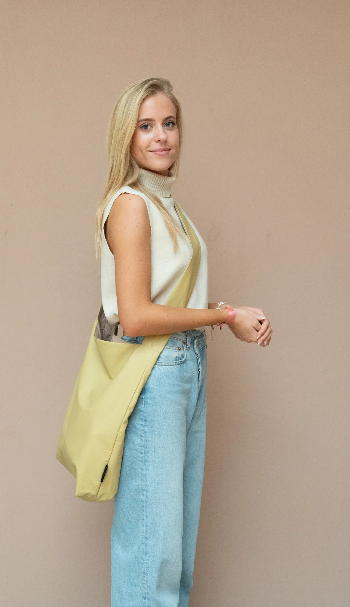 A person with long blonde hair stands against a beige wall in a sleeveless beige top and light blue jeans, carrying the Feel Good Bag - Violet Ice—showcasing modern curated style inspired by concept shop vibes. Gate21.ee