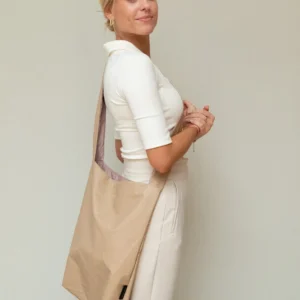 A woman with blonde hair tied back, in a white top and high-waisted trousers, stands sideways holding the Feel Good Bag - Porcini—ideal for Tallinn shopping or carrying finds from your favourite concept store. Gate21.ee