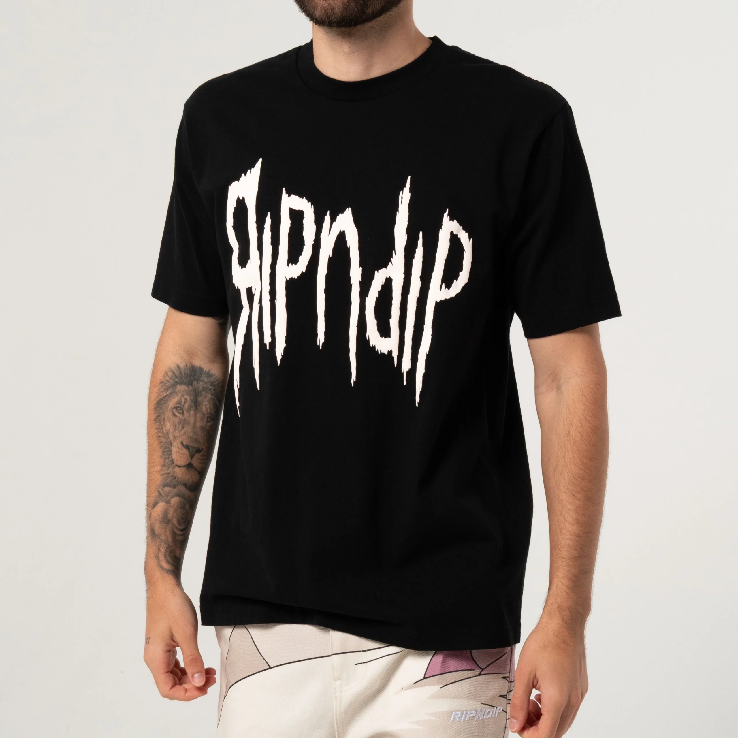 Wearing the Insano Tee Black with bold, jagged white RIPNDIP lettering, a person in light shorts—sporting a lion tattoo on their left forearm—shops at a curated concept shop. Gate21.ee