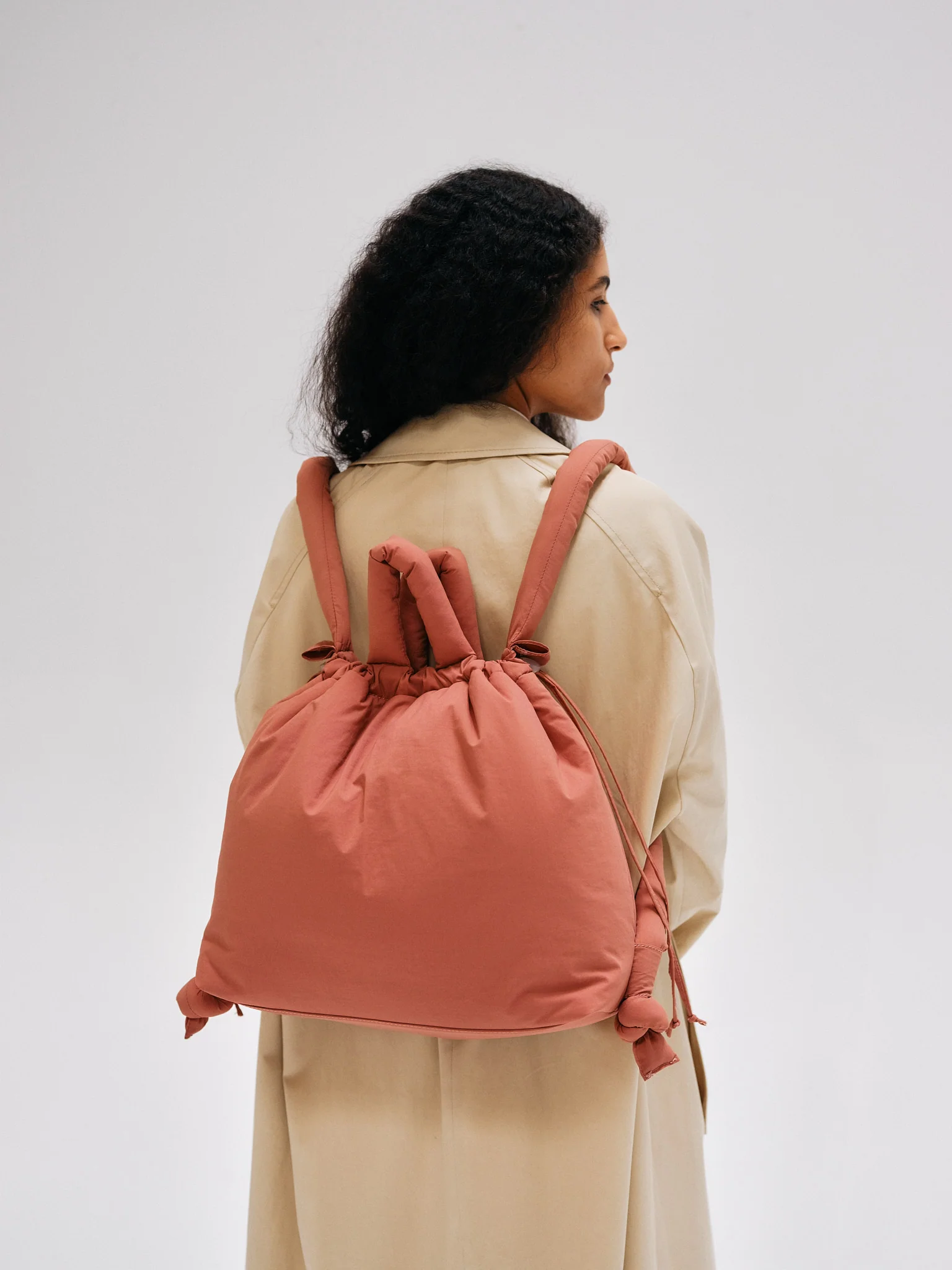 A person with long, dark curly hair in a light beige coat is seen from behind, carrying the Ona Soft Bag in Soft Peach—featuring wide padded straps—against a plain light background. Gate21.ee