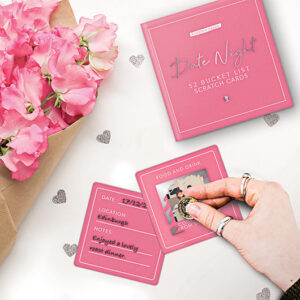 A hand scratches a pink FOOD AND DRINK card from the Date night - Scratch Cards set. Pink flowers and Estonian-designed cards are displayed on a white surface. Gate21.ee