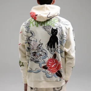 A person faces away from the camera, wearing the Travis Hoodie Off White—cream-coloured with intricate Estonian-inspired back design featuring a black cat, rose, dragon, clouds, and stylised RIPNDIP. A standout gift shop pick for unique styles. Gate21.ee
