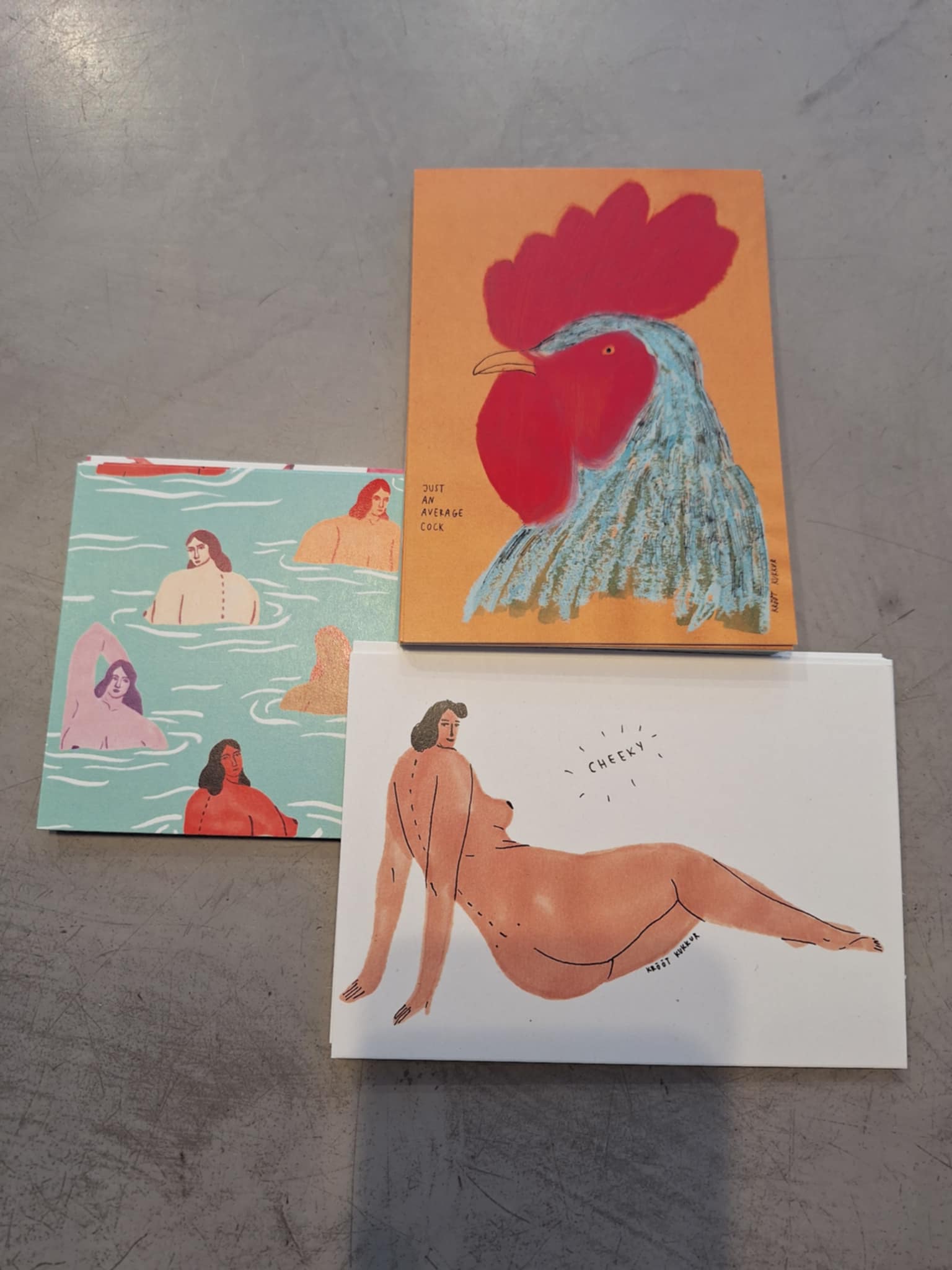 Three Krõõt Kukkur Postcards - KÕIK on a grey surface: one with a cockerel, one with swimmers, and one "cheeky" nude—showcasing the unique creativity of Estonian design. Gate21.ee