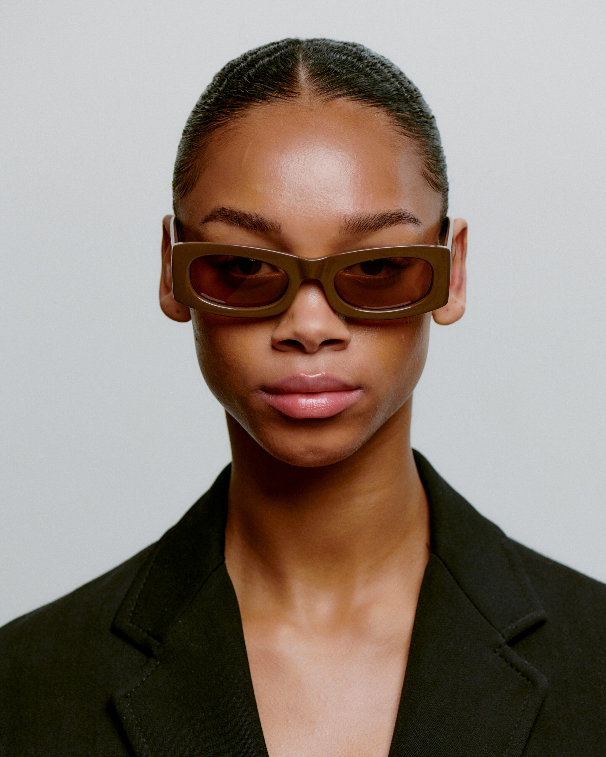 A person with smooth, medium-brown skin and slicked-back hair models the FRIDA - Chocolate rectangular sunglasses with a black blazer against a light background—a style perfect for any curated concept shop or Tallinn shopping scene. Gate21.ee
