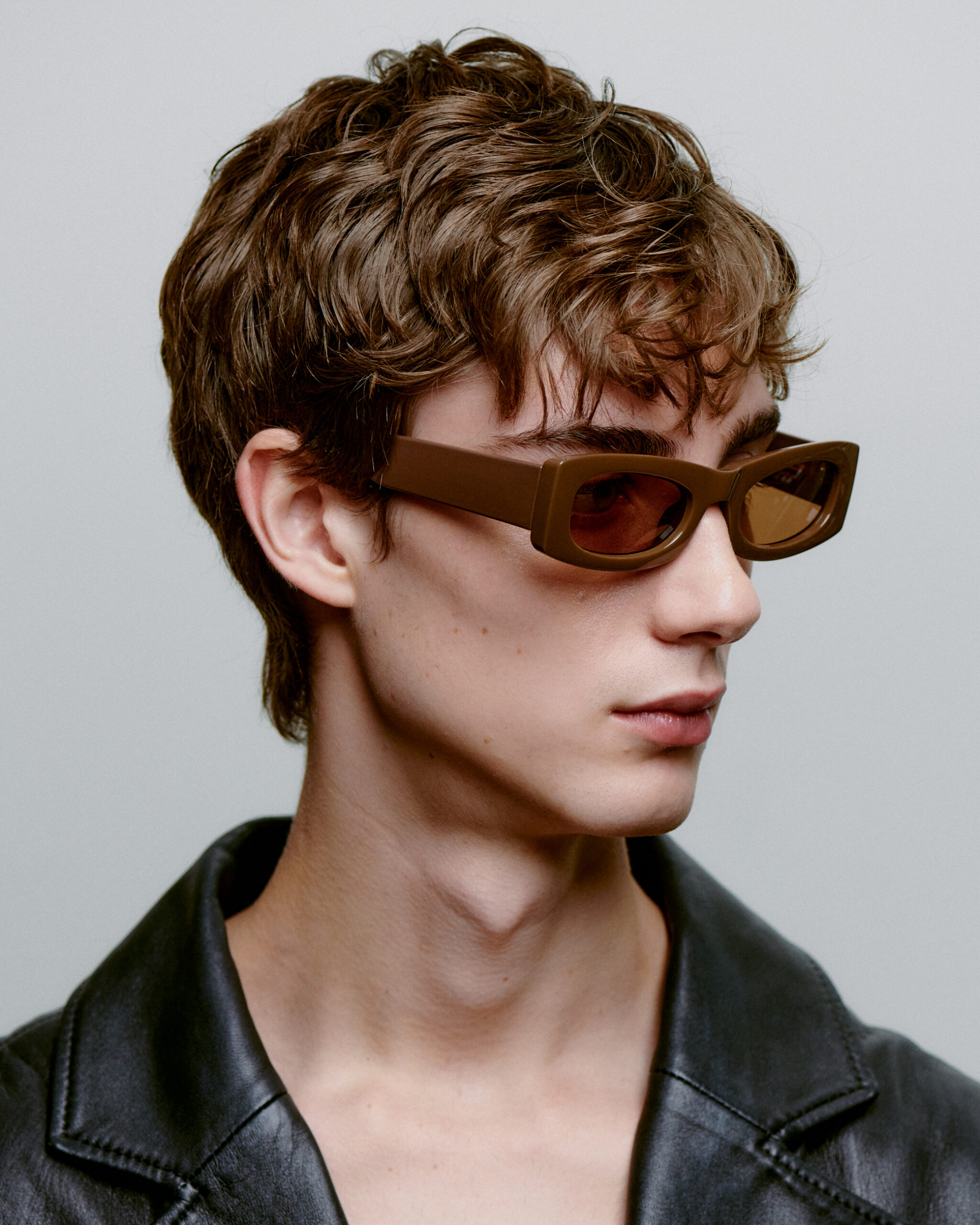 A young person with short, wavy brown hair wears FRIDA - Chocolate rectangular sunglasses and a black leather jacket, looking left against a plain light background—an effortless style inspired by Tallinn shopping and Estonian design. Gate21.ee