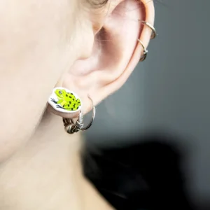 A close-up of a person’s ear wearing Konnad Kõrvarõngad—silver hoop earrings featuring a yellow and green frog charm inspired by Estonian design. A smaller hoop accents the upper ear, with a blurred background for an artsy Tallinn vibe. Gate21.ee