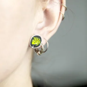 Close-up of a person’s ear wearing three silver hoops and Konnad lehekesel Kõrvarõngad, featuring a round black and yellow design. Perfect for those seeking unique ideas at a concept shop. Blurred neutral background. Gate21.ee