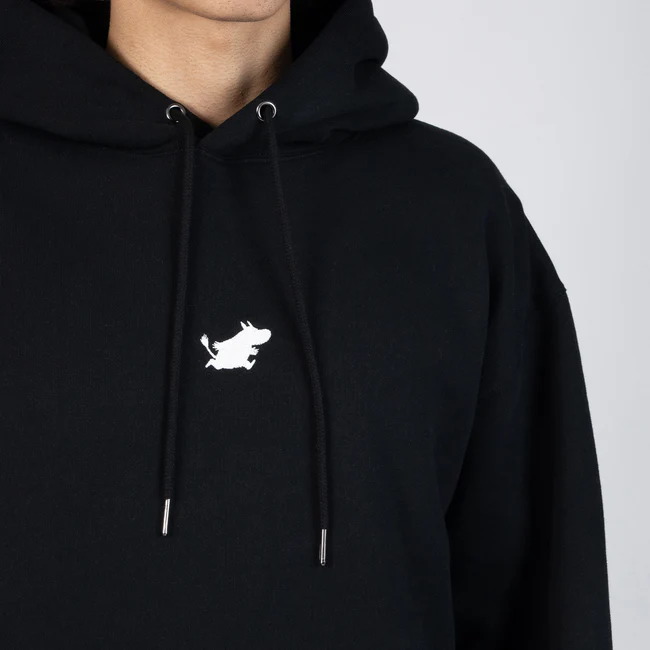 A person models the Moomin Hoodie - Black, which features a small white embroidered cartoon bull on the front. Only their lower face, neck, and upper torso are visible. Gate21.ee