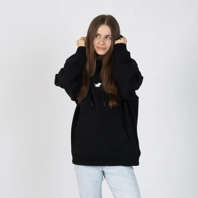 A person with long brown hair wears the Moomin Hoodie - Black, featuring a small white chest graphic, paired with light blue jeans. Standing against a white background, they hold the hood strings and gaze slightly to the side. Gate21.ee