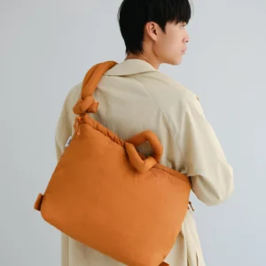 A person with short dark hair in a beige trench coat stands sideways, carrying the large, orange Ona Soft Bag - Oxide—an eye-catching Estonian design—over their shoulder against a plain, minimalistic background. Gate21.ee