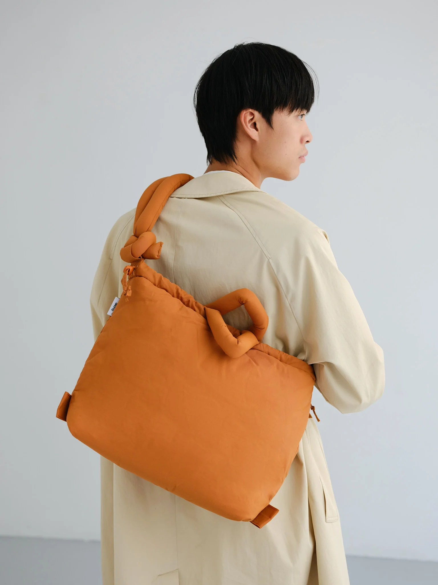 A person with short dark hair in a beige trench coat stands sideways, carrying the large, orange Ona Soft Bag - Oxide—an eye-catching Estonian design—over their shoulder against a plain, minimalistic background. Gate21.ee