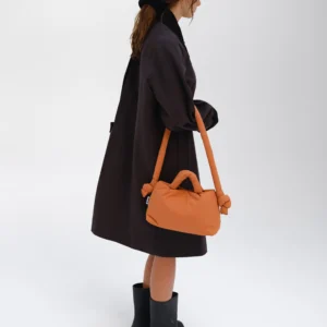 A person stands against a white background, wearing a loose dark coat and black boots, carrying the Mini Ona Soft Bag - Oxide with its signature knotted handles—a striking example of modern Estonian design. Their hair is down, and they face slightly away from the camera. Gate21.ee