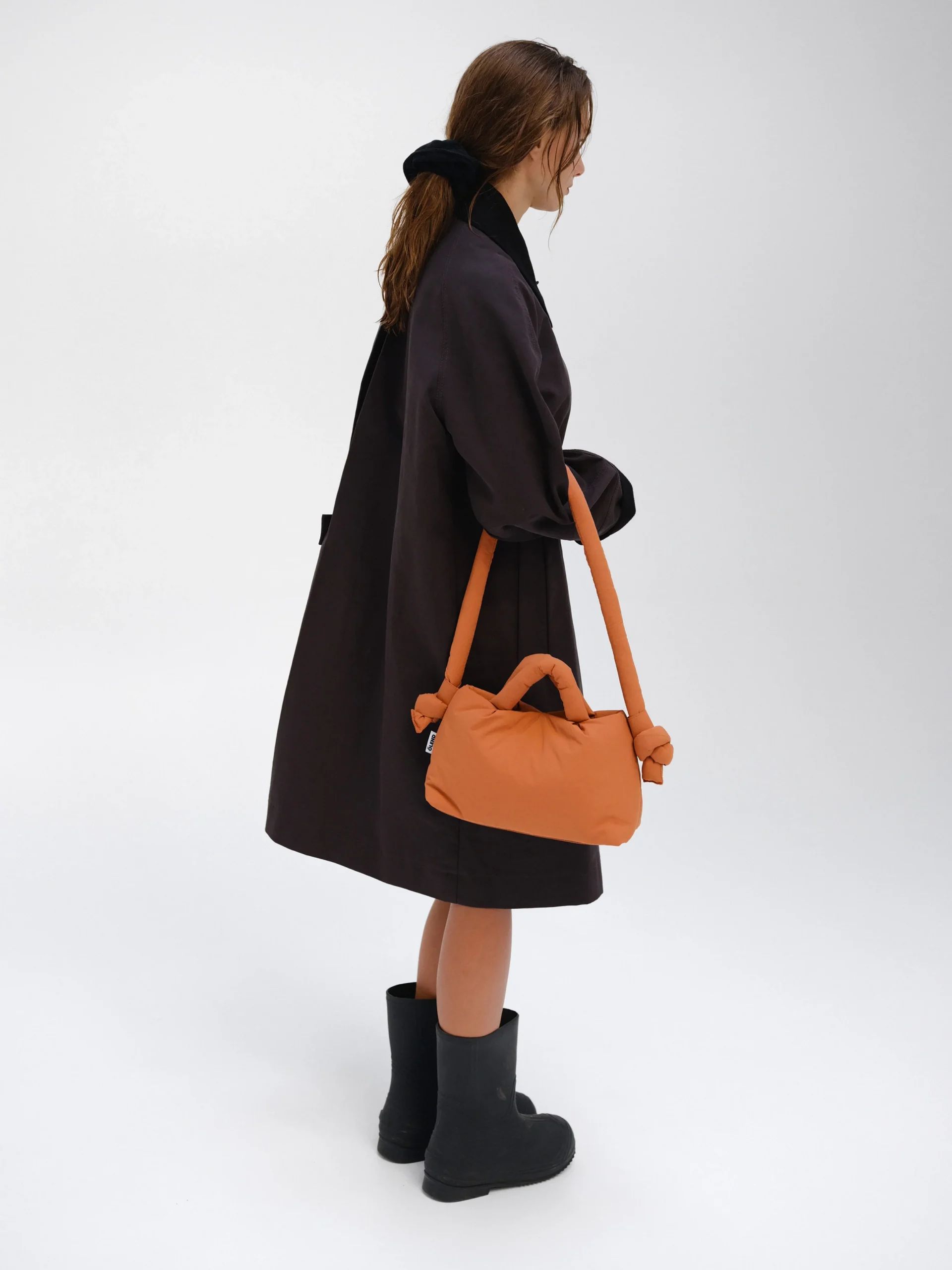 A person stands against a white background, wearing a loose dark coat and black boots, carrying the Mini Ona Soft Bag - Oxide with its signature knotted handles—a striking example of modern Estonian design. Their hair is down, and they face slightly away from the camera. Gate21.ee