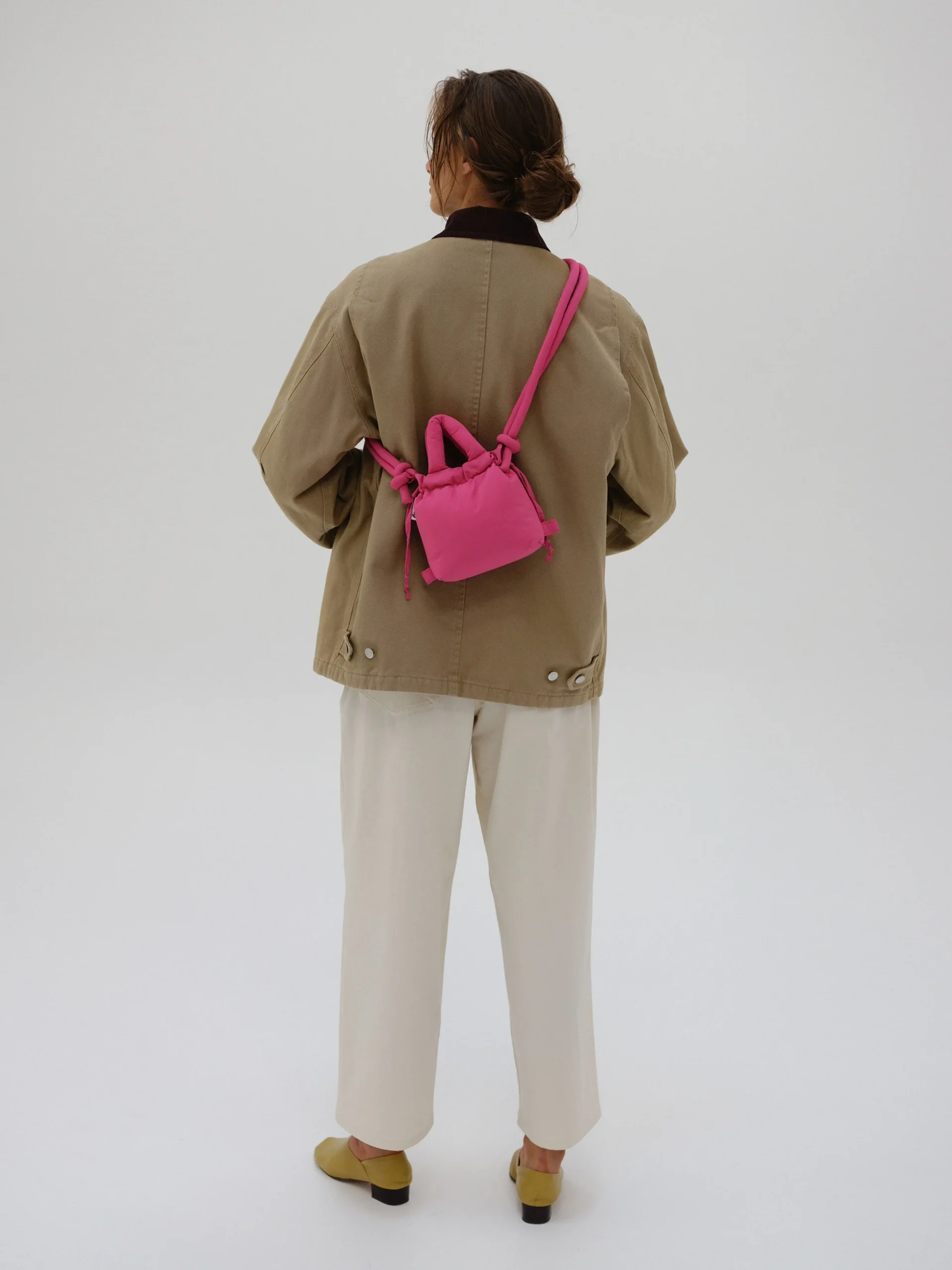 A person stands facing away from the camera, wearing neutral-toned clothing and carrying the Micro Ona Soft Bag - Pink—an eye-catching accessory for exploring new ideas at a concept store. The background is plain white. Gate21.ee