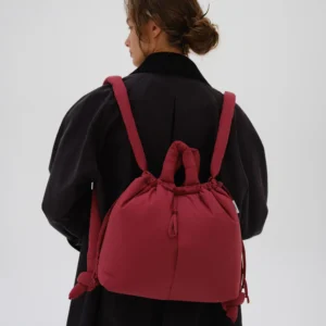 A person in a dark coat stands with their back to the camera, carrying the Ona Soft Bag in Cherry Red—its knotted handle and bold colour make it look like a piece from a curated concept store. The background is plain and light-coloured. Gate21.ee