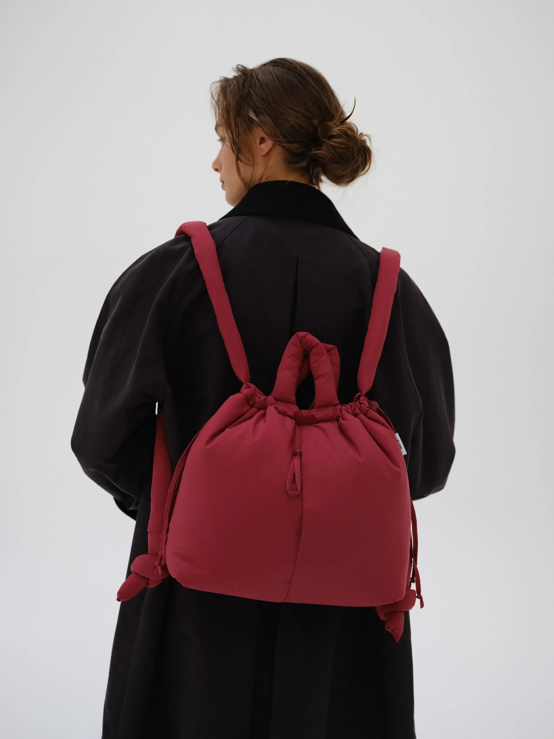 A person in a dark coat stands with their back to the camera, carrying the Ona Soft Bag in Cherry Red—its knotted handle and bold colour make it look like a piece from a curated concept store. The background is plain and light-coloured. Gate21.ee