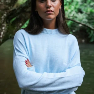 A woman with straight brown hair stands by a river and trees, wearing the Jumper - Baby Blue with arms crossed and looking at the camera—ideal for anyone seeking unique style ideas or Tallinn shopping inspiration. Gate21.ee