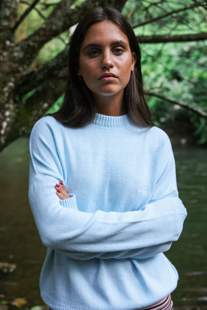 A woman with straight brown hair stands by a river and trees, wearing the Jumper - Baby Blue with arms crossed and looking at the camera—ideal for anyone seeking unique style ideas or Tallinn shopping inspiration. Gate21.ee