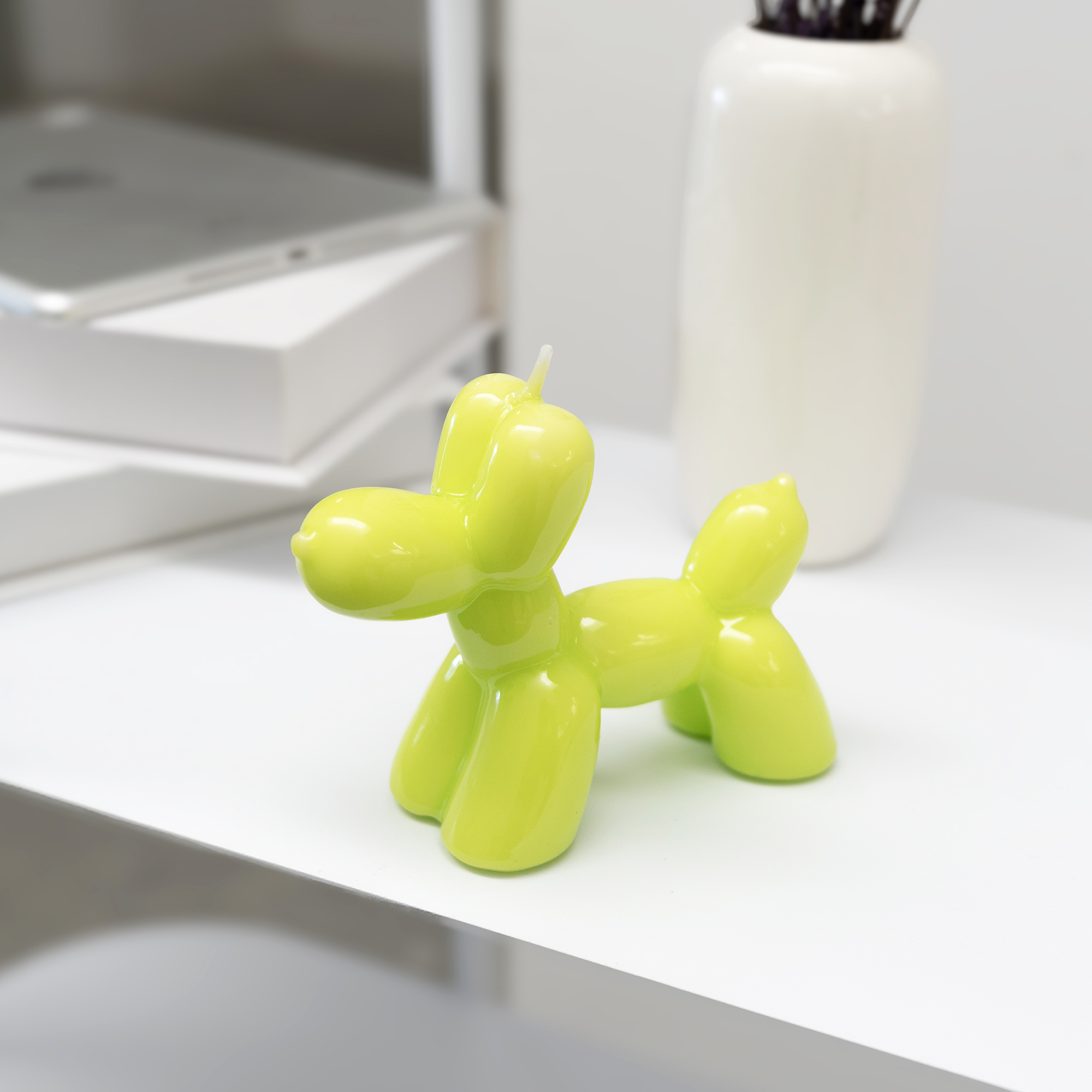 The Balloon Dog Candle - Lime adds playful charm to modern, minimal spaces as it sits on a white tabletop near books and a vase—an eye-catching decor piece, ideal for gift shops. Gate21.ee