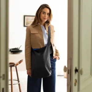 A woman stands indoors by an open door, wearing a tan jacket, blue shirt, dark wide-leg trousers, and carrying the Feel Good Bag in Midnight Blue—embodying curated Tallinn style. A kitchen with stools and a framed picture can be seen behind her. Gate21.ee