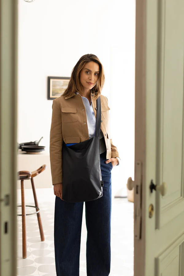 A woman stands indoors by an open door, wearing a tan jacket, blue shirt, dark wide-leg trousers, and carrying the Feel Good Bag in Midnight Blue&mdash;embodying curated Tallinn style. A kitchen with stools and a framed picture can be seen behind her. Gate21.ee