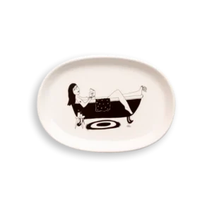 The "How to Relax - Tray" is a white oval plate with a black line drawing of a woman in a bath, drink and book in hand—an Estonian design gem perfect for anyone looking for unique Tallinn shopping finds. Gate21.ee
