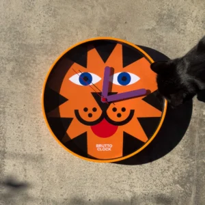 The TIIGER Wall Clock features a round face with a stylised orange lion design, the words BRUTTO CLOCK, and part of a black cat’s head at the edge. Displaying about 10:15, it’s curated for concept shops and set on a concrete surface. Gate21.ee