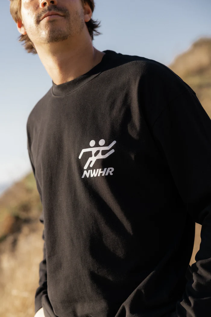 A person wears the NWHR TO GO T-shirt - Black, featuring a white graphic and NWHR lettering on the chest, standing outdoors in natural light—an ideal pick for fans of Estonian design or those seeking unique concept shop finds. Gate21.ee