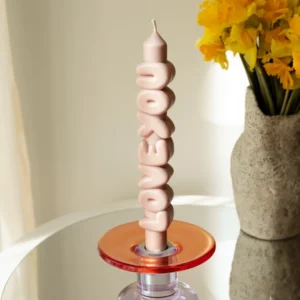 The Love You 3D Candle - Cream, featuring a sculptural wavy design and curated Estonian style, sits in a red and purple glass holder on a reflective surface. In the background, there's a stone vase filled with yellow flowers. Gate21.ee