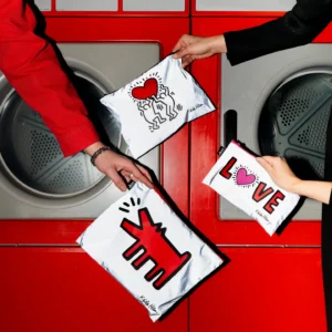 Two hands hold the Keith Haring 3x Zip Pockets LOQI Reflective pouches, each with a reflective zip pocket design—one showcases a red barking dog, another displays a radiating red heart, and the third spells LOVE with a heart as the O—in front of red washing machines. Gate21.ee